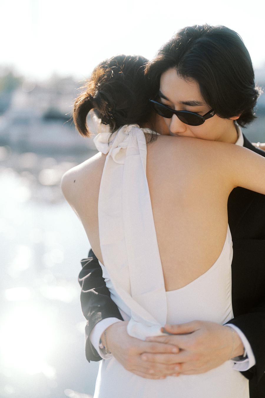 Couple embracing closely by a bright marina, one in a white dress, the other in a black suit and sunglasses, with sunlight reflecting on the water.