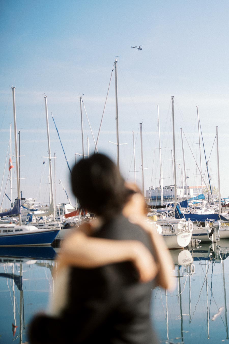Couple embracing in soft focus with sailboats and marina in the background under a clear sky.