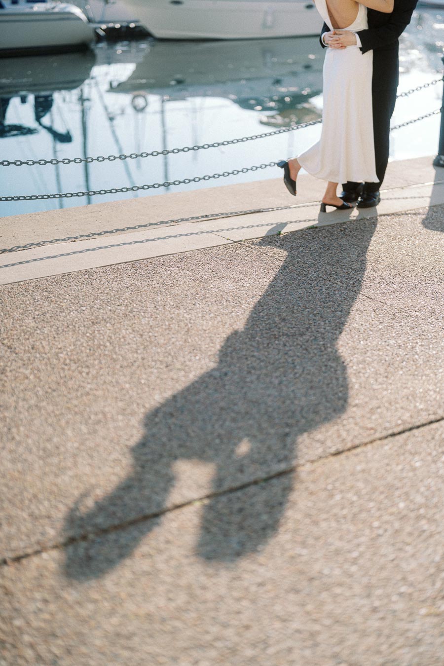 Couple in formal attire embracing by the waterfront, with their shadow clearly visible on the pavement and boats in the background.