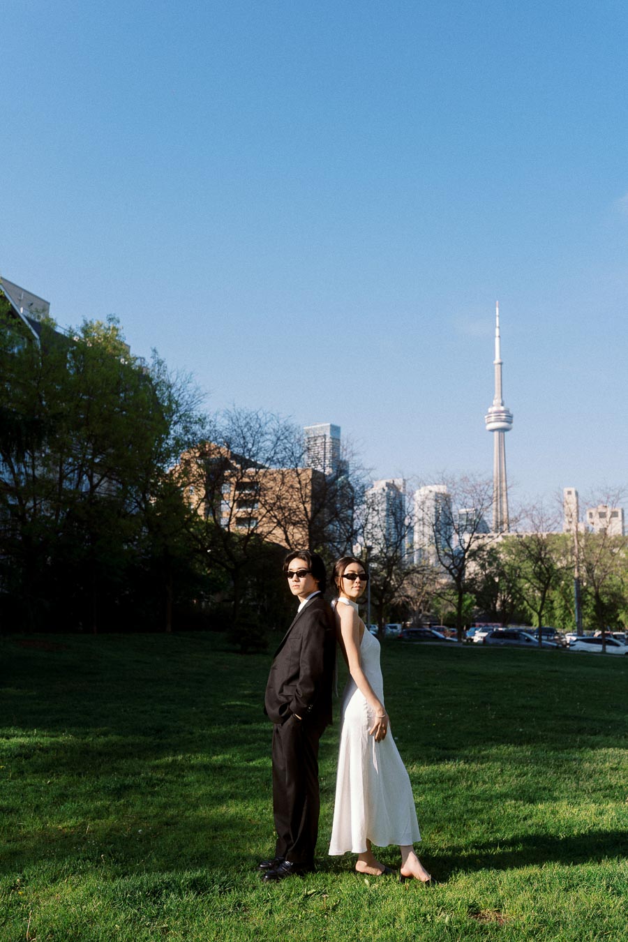 Couple standing back to back on green grass, both in formal attire and sunglasses, with Toronto CN Tower and city skyline in the background.