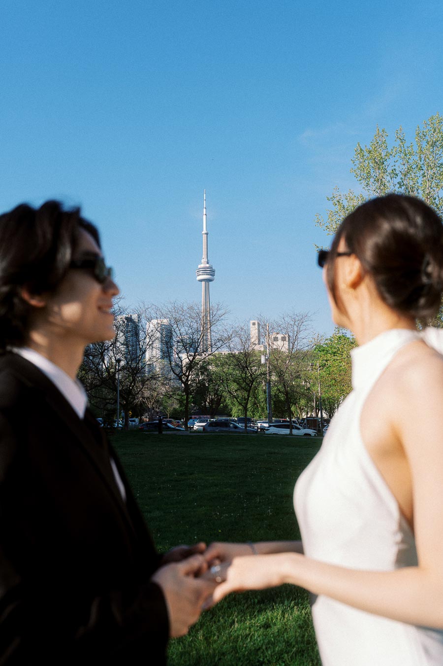 Couple in formal attire holding hands in a park, CN Tower and Toronto skyline in the background on a sunny day.