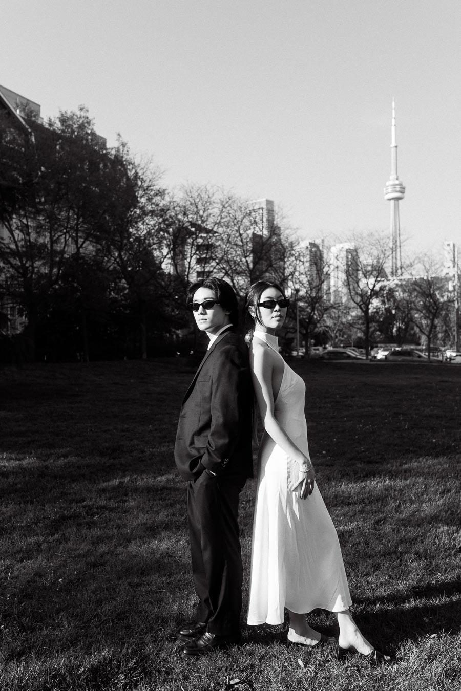 Couple standing back to back on grass, both in sunglasses, with Toronto skyline and CN Tower visible behind them.