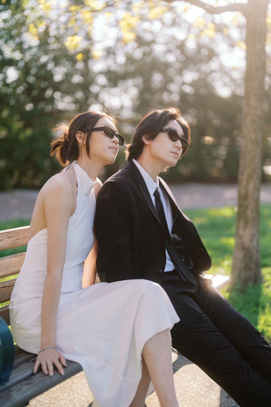 A stylish couple in sunglasses, one in a white dress and the other in a black suit, sitting together on a park bench in soft evening light.