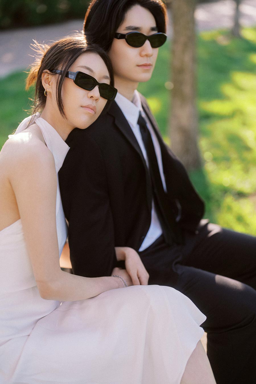 Couple sitting together on a park bench, both wearing sunglasses, with one in a white dress and the other in a black suit, in soft afternoon light.