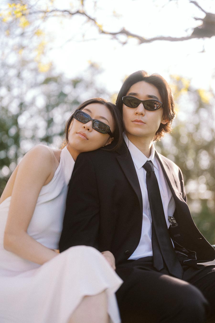 Couple in formal outfits and sunglasses, sitting close together outdoors with soft sunlight and blurred trees in the background.