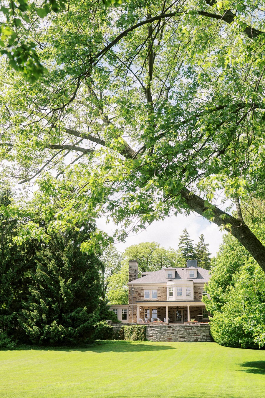 Historic stone mansion seen beyond green lawn and leafy tree branches in summer sunlight.
