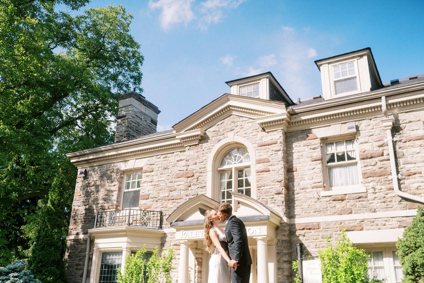 Engaged couple kissing in front of stone mansion entrance on a sunny day.