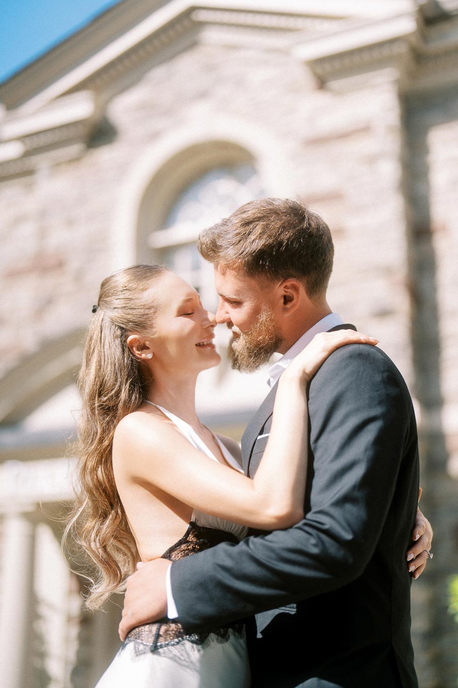 Anastasiia and Petr embracing closely in front of Paletta Mansion's stone facade on a sunny day.
