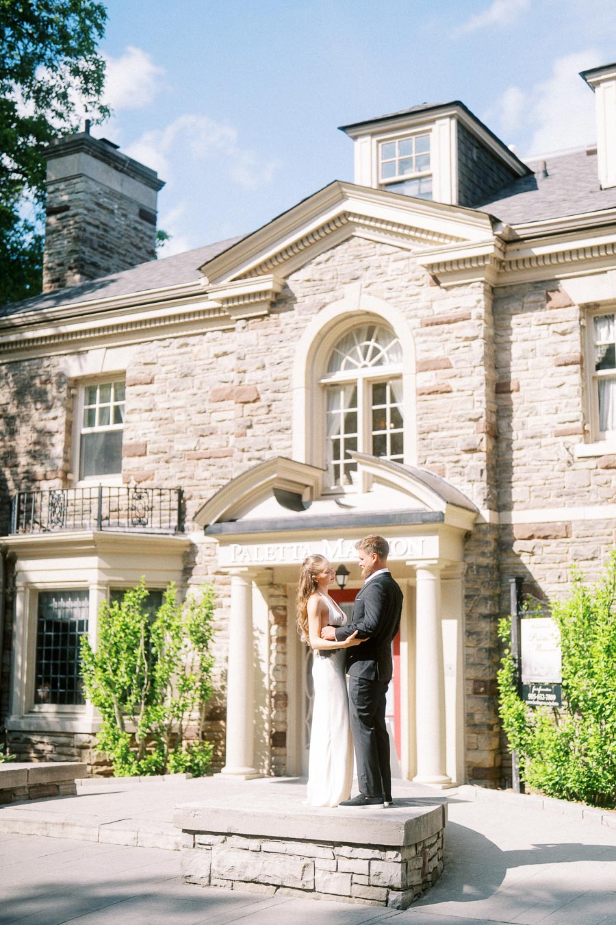 Engaged couple facing each other on stone platform in front of Paletta Mansion entrance, surrounded by greenery.