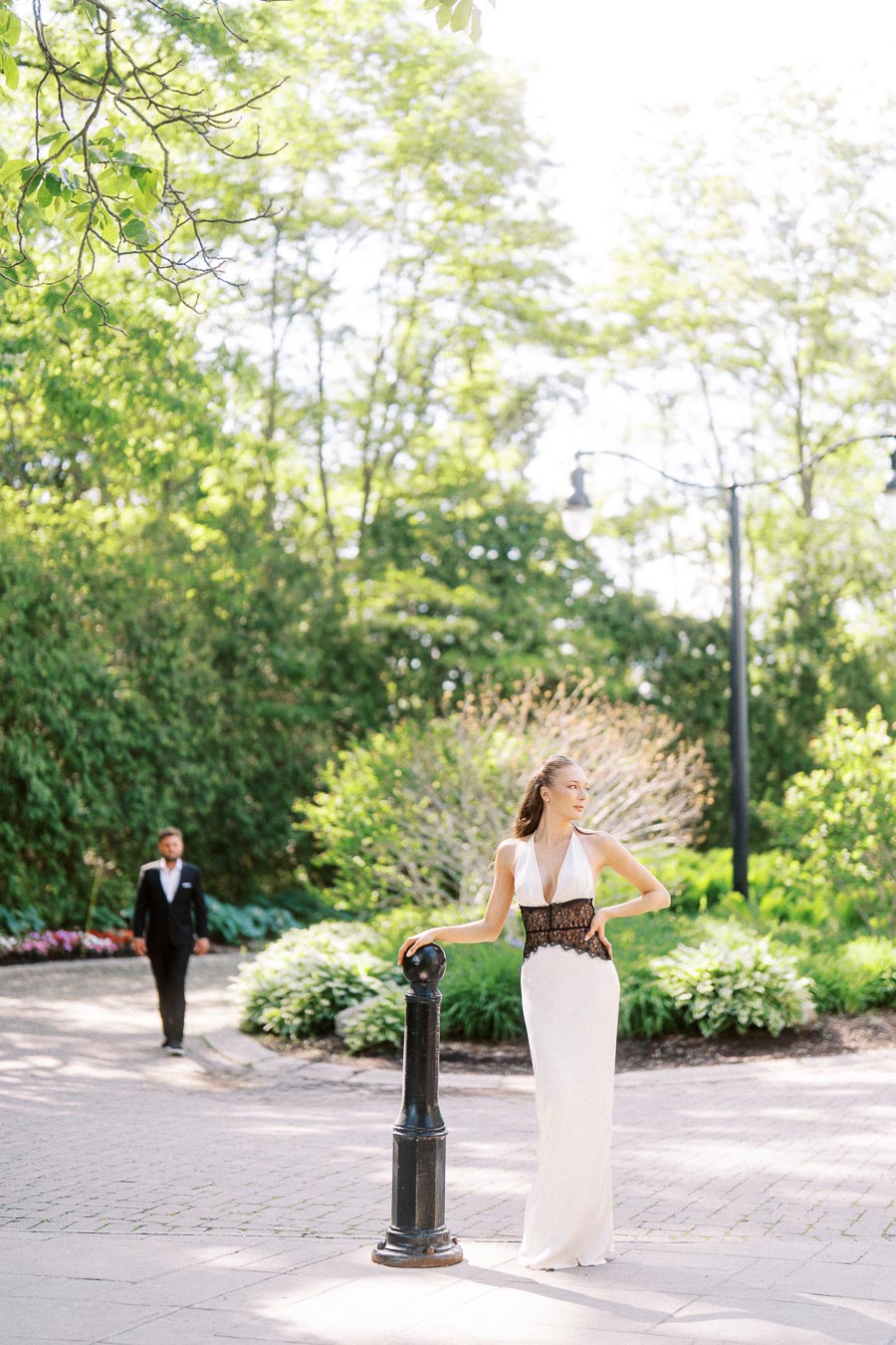 Woman in a white dress stands by a black post on a garden path, man in suit walking behind among greenery.