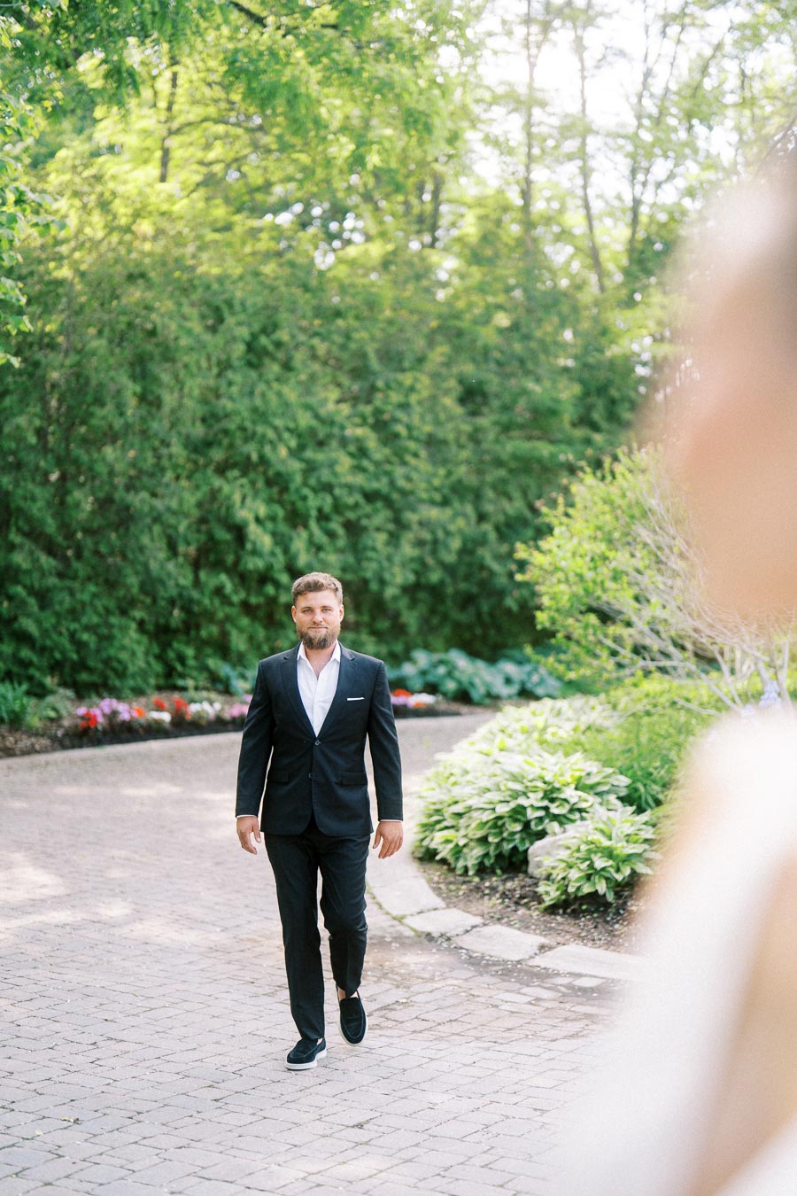 Man in dark suit walking on stone path through garden with blurred figure in foreground.