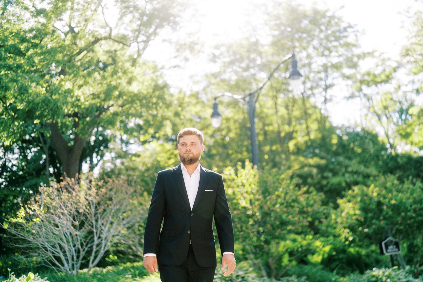 Man in a dark suit walking in a sunlit garden with trees and greenery in the background.