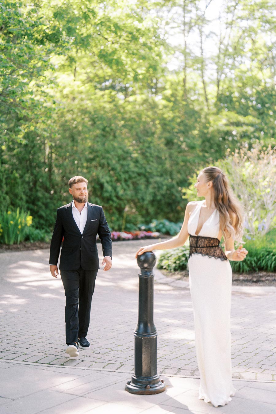 Anastasiia in a white dress stands near a post as Petr approaches on a garden path lined with greenery.