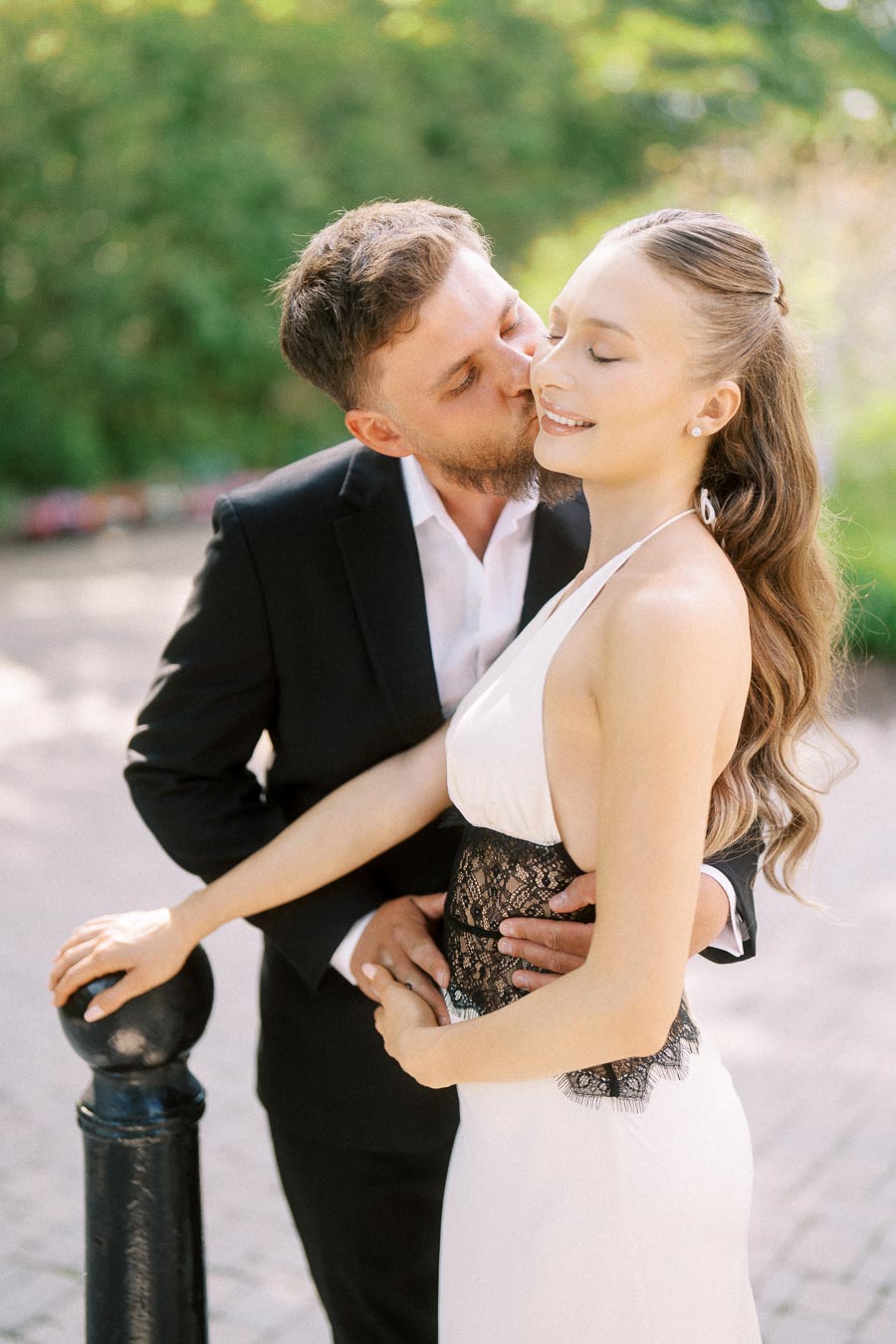 Engaged couple standing together, Petr kissing Anastasiia's cheek, sunlit garden background.