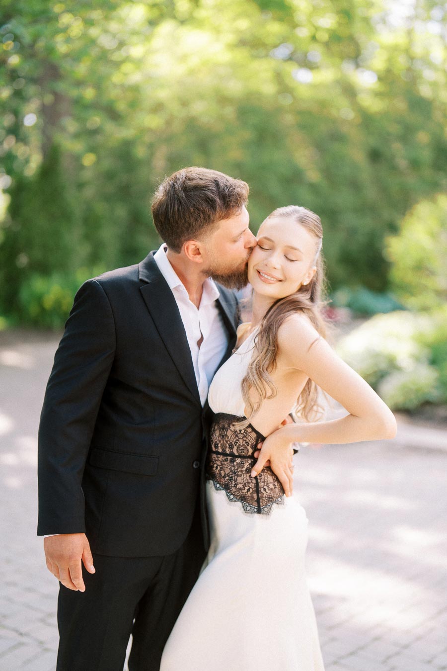 Engaged couple embracing on a sunlit garden path, man kissing woman's cheek, both dressed formally.