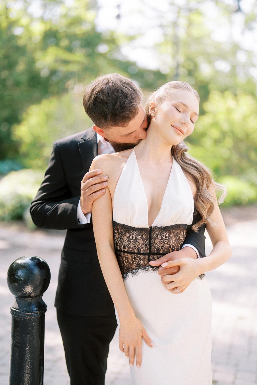 Engaged couple embracing outdoors, man kissing woman's neck, soft sunlight and greenery behind them.