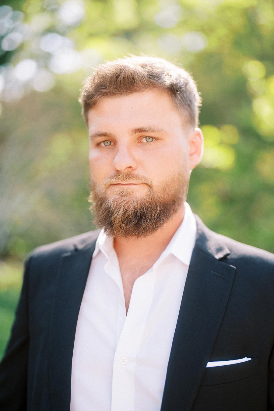 Man with beard in dark suit jacket and white shirt outdoors, greenery in background.