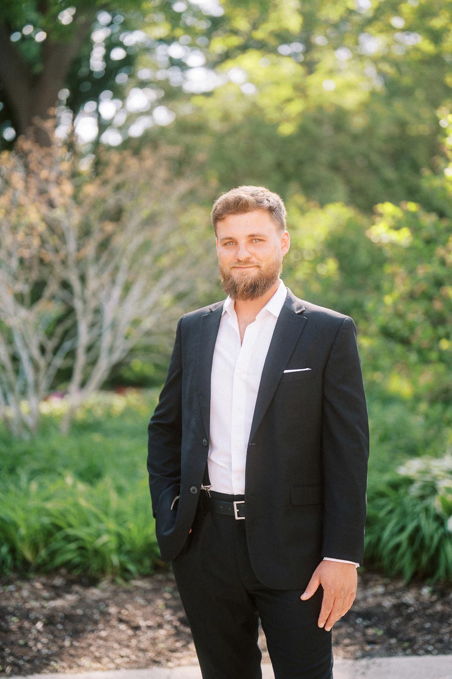 Bearded man in dark suit and white shirt standing in sunlit garden with trees and plants.