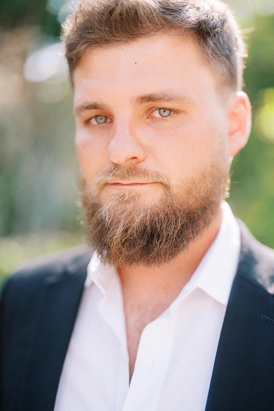 Bearded man in white shirt and dark jacket standing outdoors in soft sunlight.