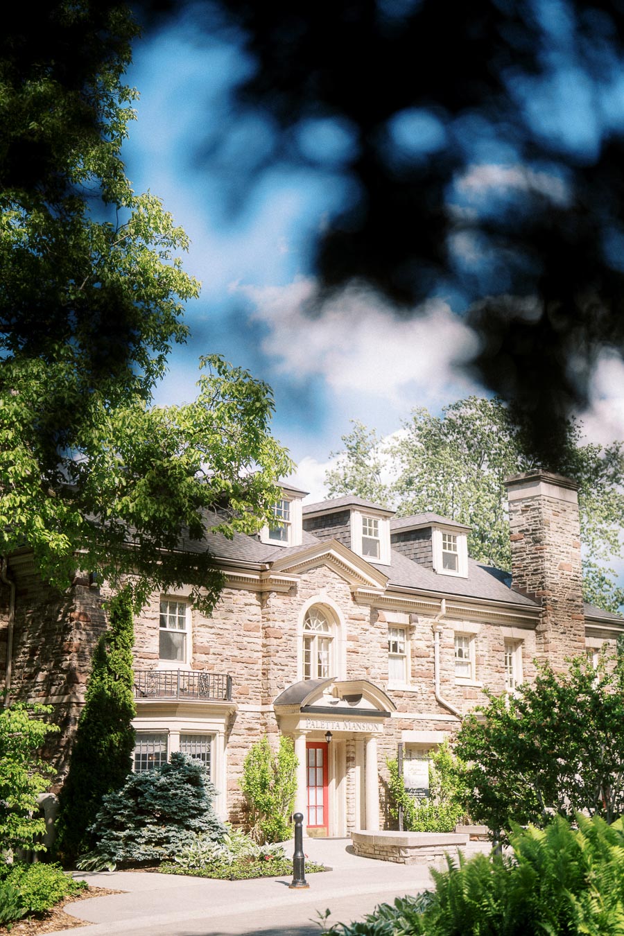 Paletta Mansion stone facade framed by leafy branches and shrubs under blue sky.
