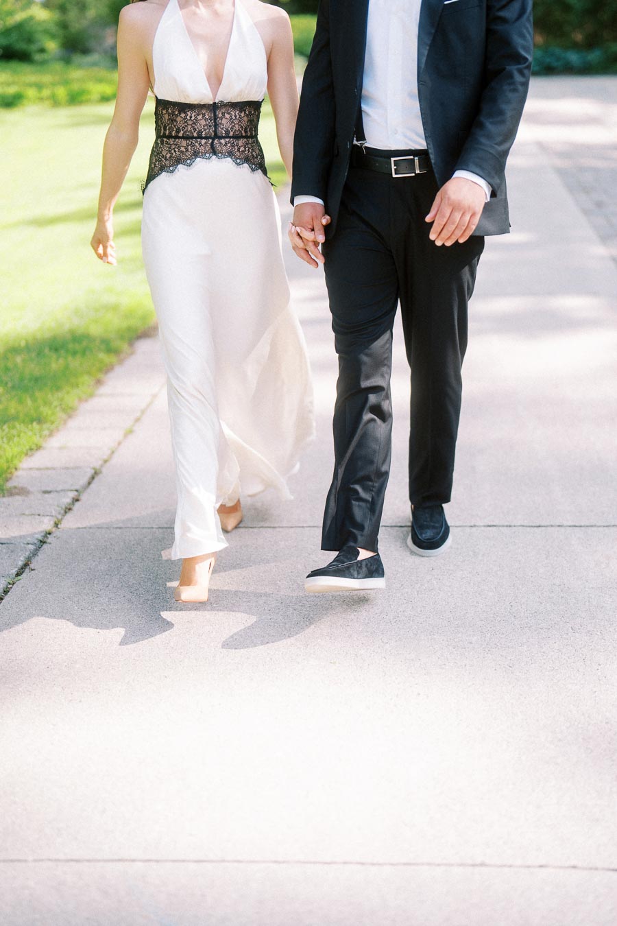 Engaged couple holding hands, walking on a paved path in formal attire, sunlit surroundings.