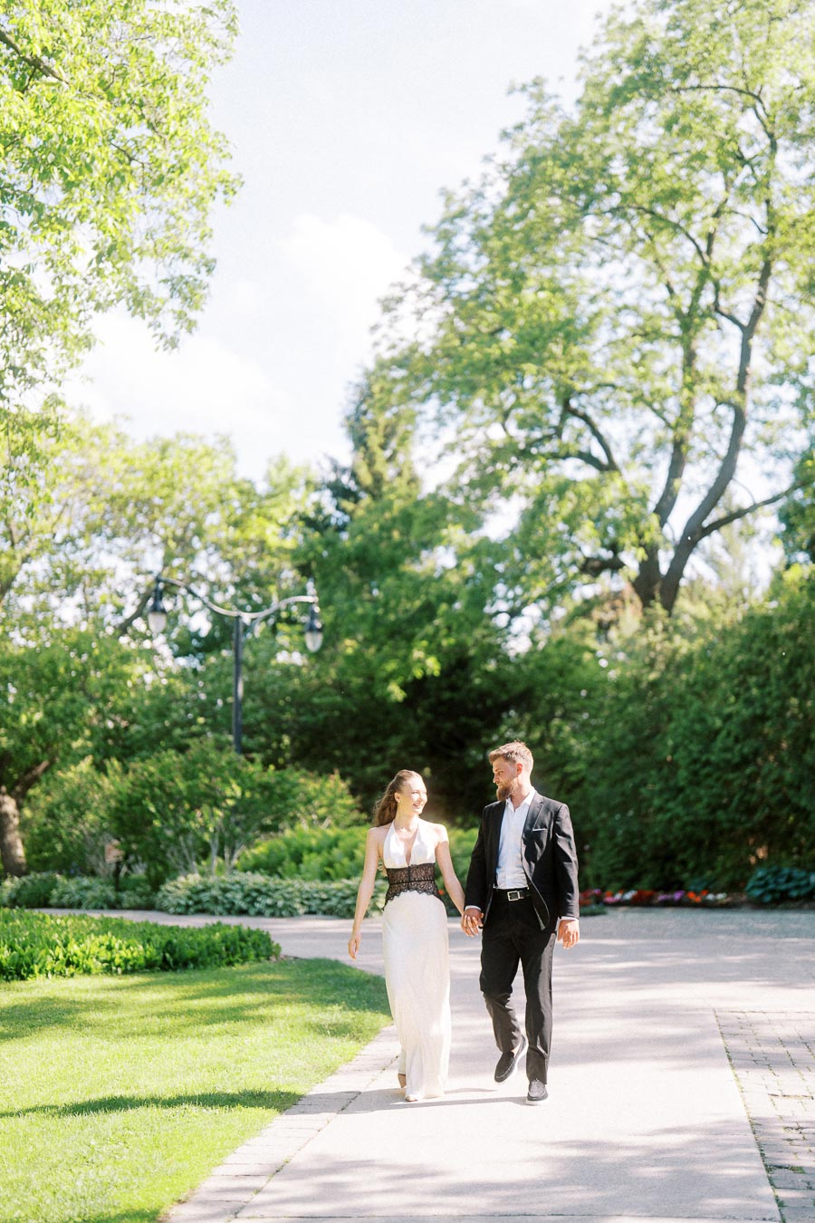 Anastasiia and Petr walk hand-in-hand on a sunny garden path surrounded by green trees.
