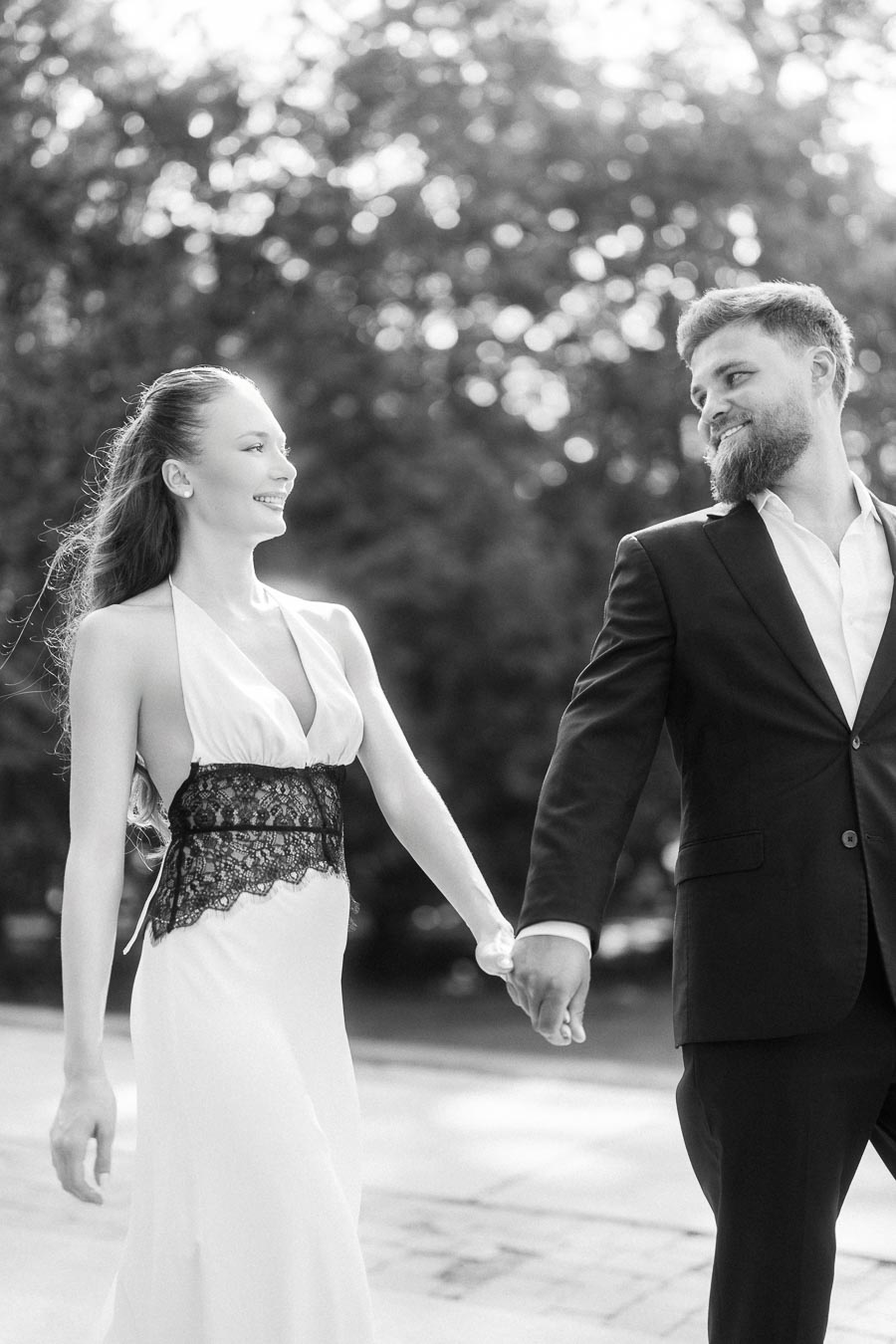 Anastasiia and Petr holding hands and smiling at each other in black and white outdoors.