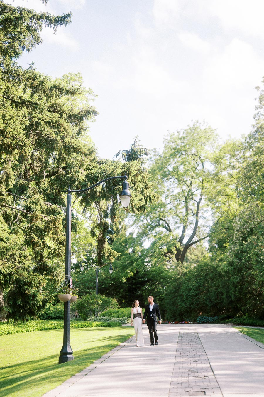 Engaged couple walking on a wide garden path bordered by green lawns and tall trees under a bright sky.