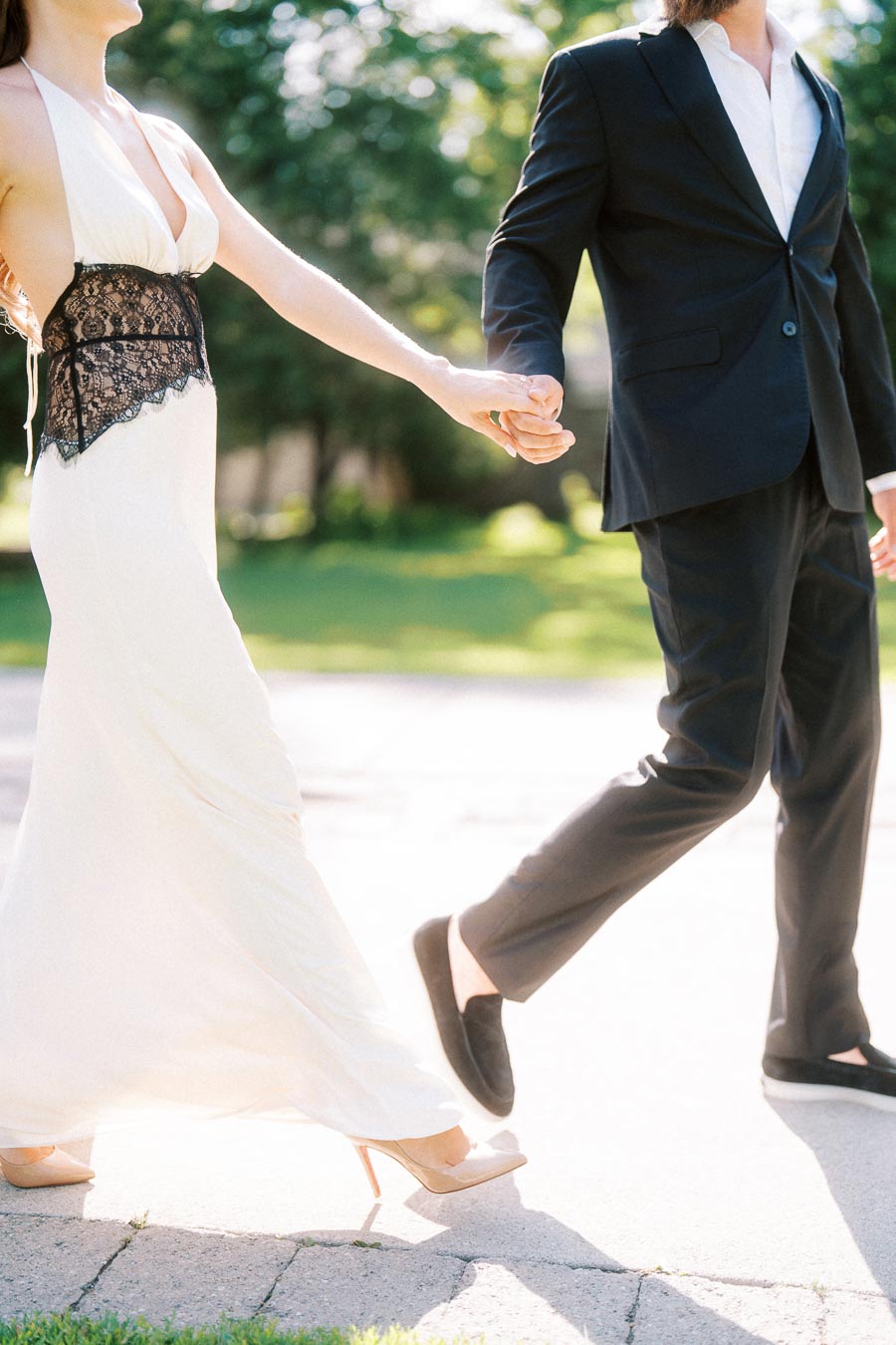 Engaged couple holding hands while walking on a bright garden path, only lower bodies visible.