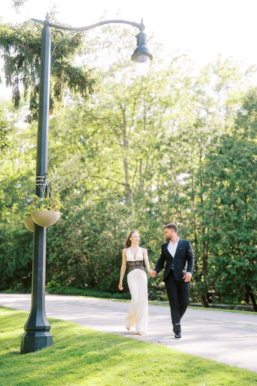 Anastasiia and Petr hold hands while walking on a paved path near a lamppost and green trees.