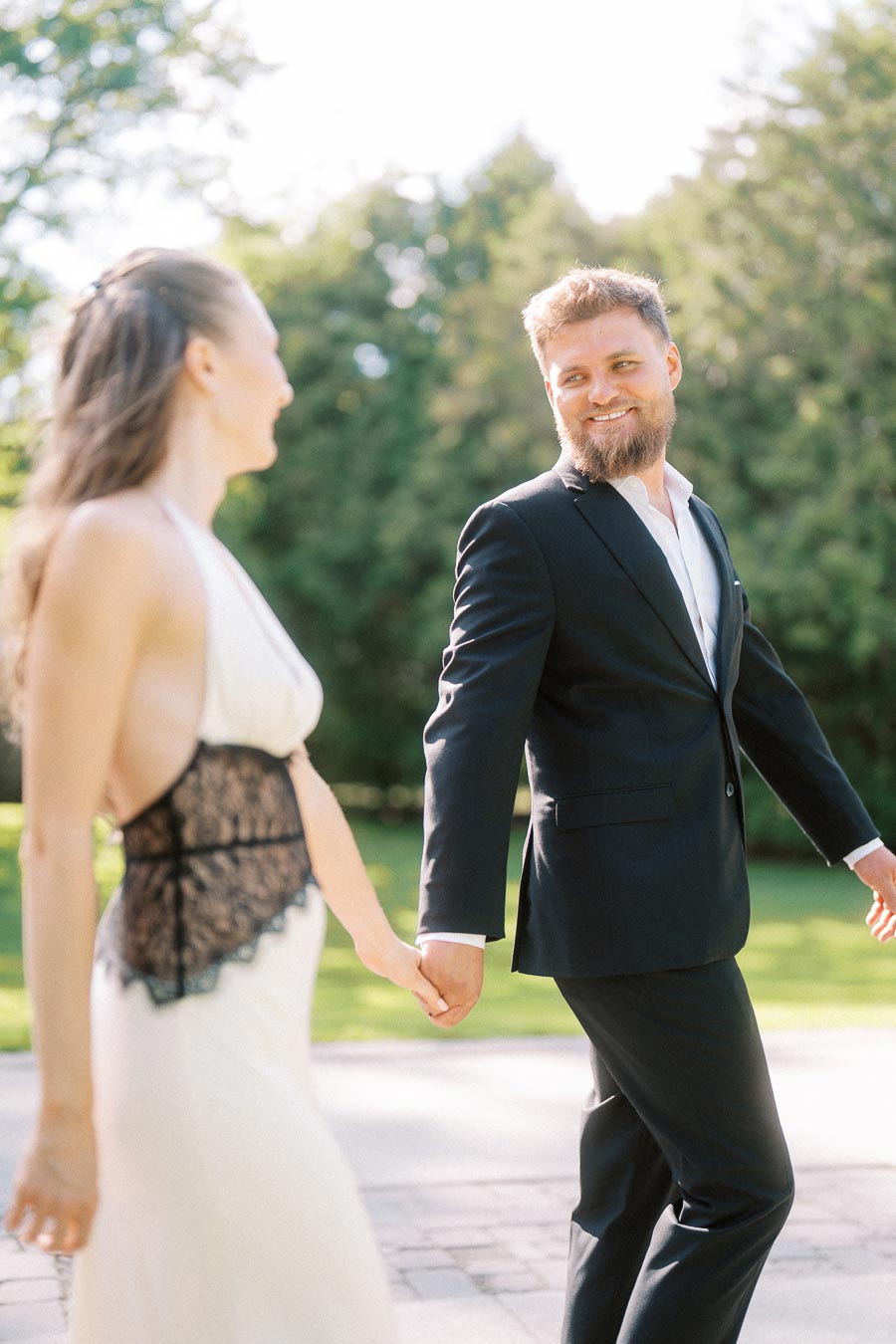 Engaged couple walking hand in hand on a sunlit path with trees in the background.