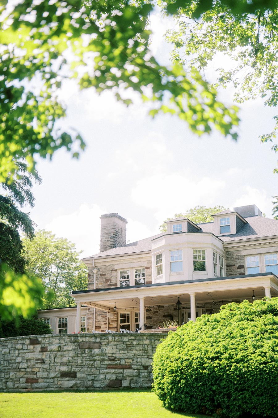 Paletta Mansion exterior with stone walls and large windows, leafy branches overhead, bright summer light.