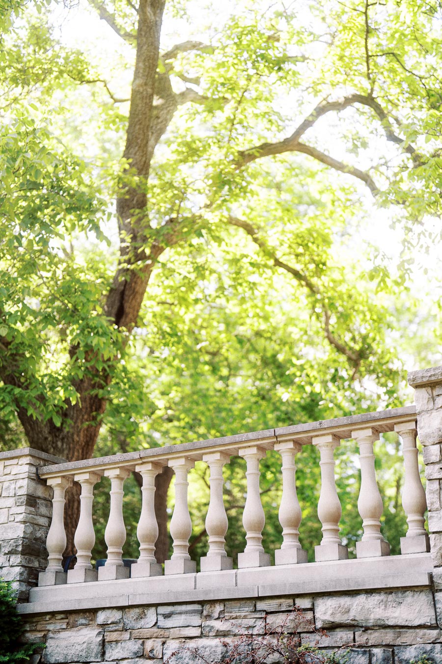 Stone balustrade with decorative columns in front of leafy green trees and sunlight.