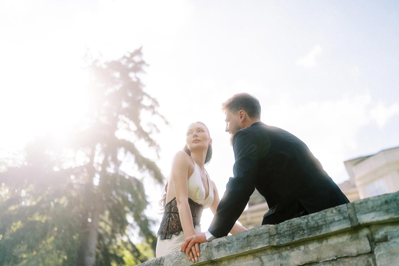 Anastasiia and Petr leaning on stone railing, sunlight streaming, trees and mansion in background.