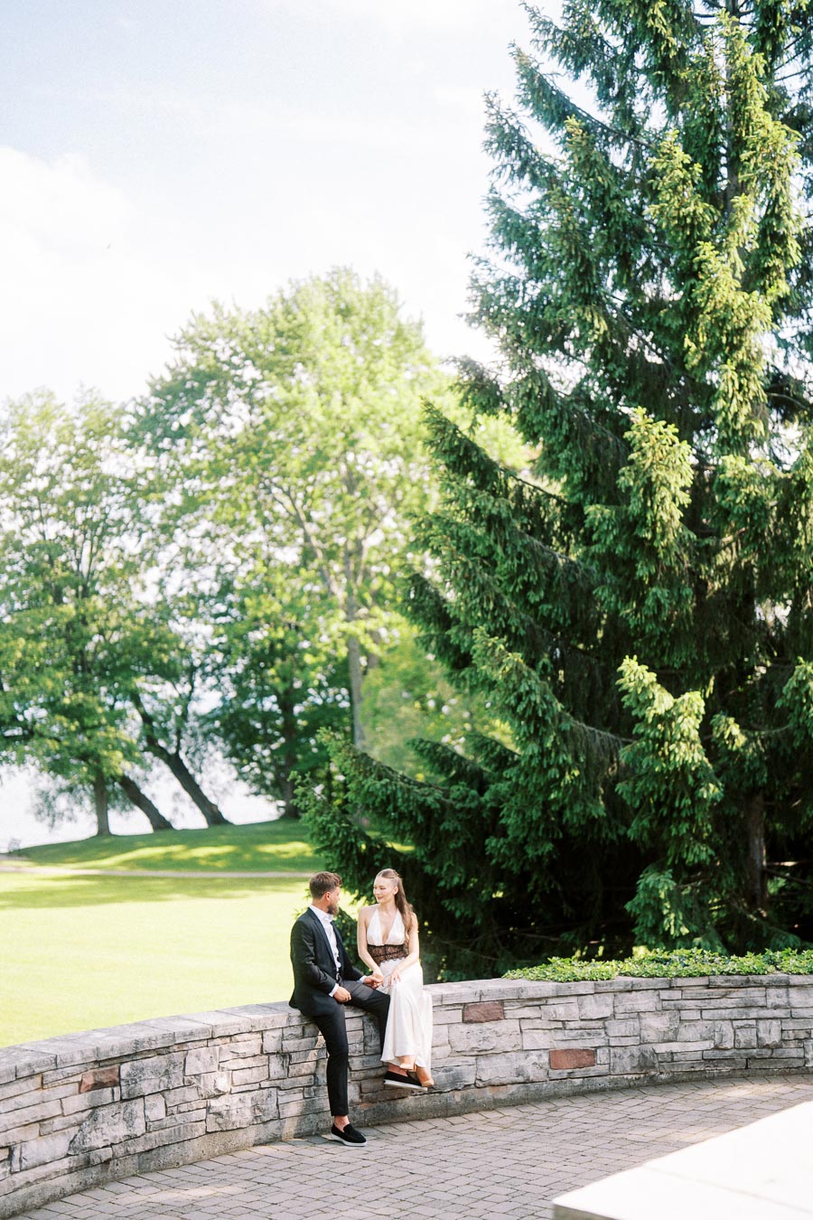 Anastasiia and Petr seated on a curved stone wall with trees and lawn in the background.