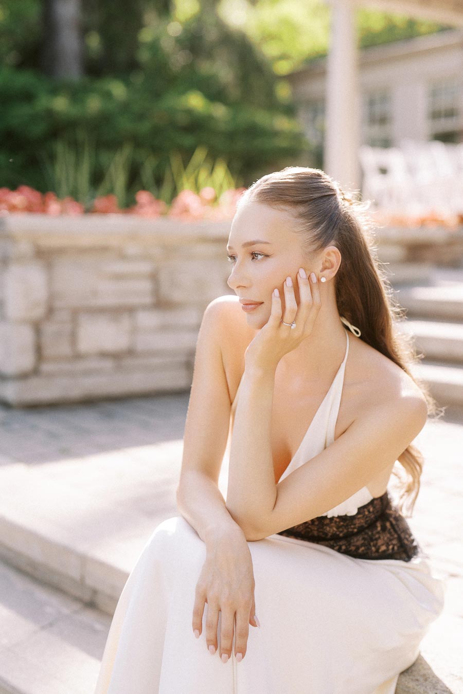 Anastasiia seated on stone steps, hand on chin, engagement ring visible, garden background.