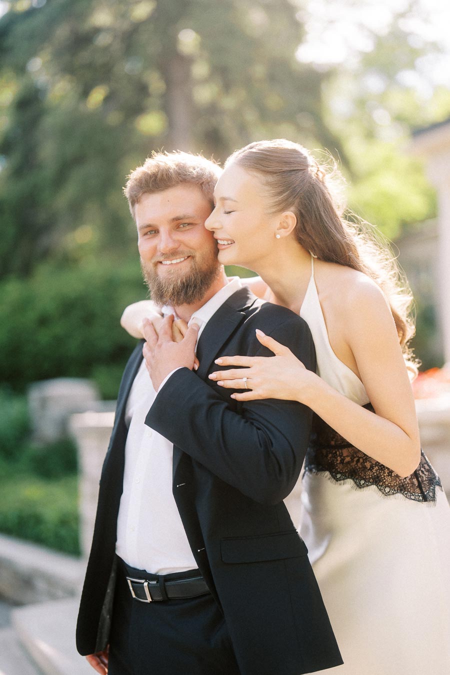 Anastasiia hugs Petr from behind, engagement ring visible, both smiling in outdoor sunlight.