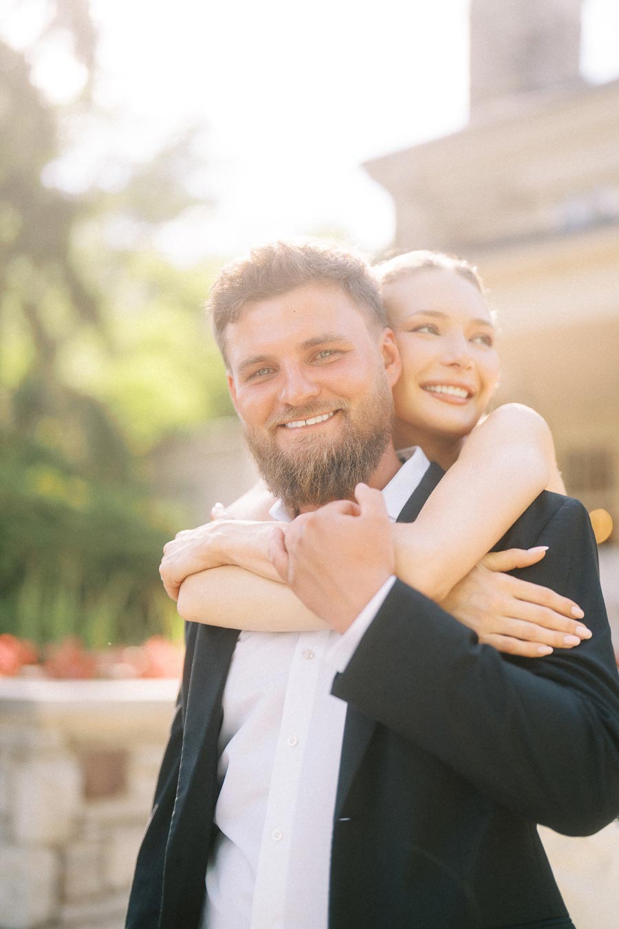 Anastasiia hugging Petr from behind outdoors with soft sunlight and blurred greenery in background.