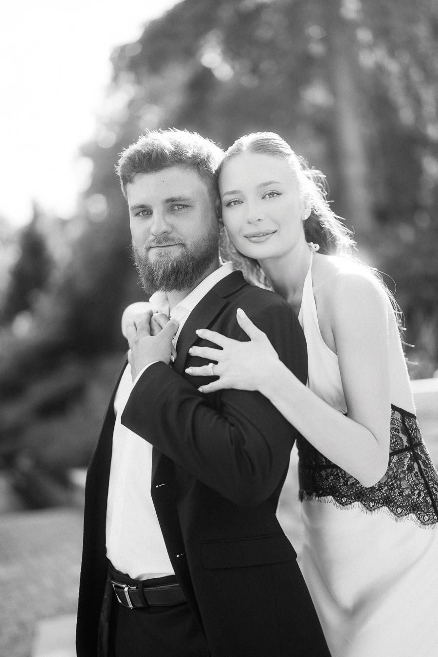 Anastasiia embracing Petr from behind, both facing the camera in black and white outdoor portrait.