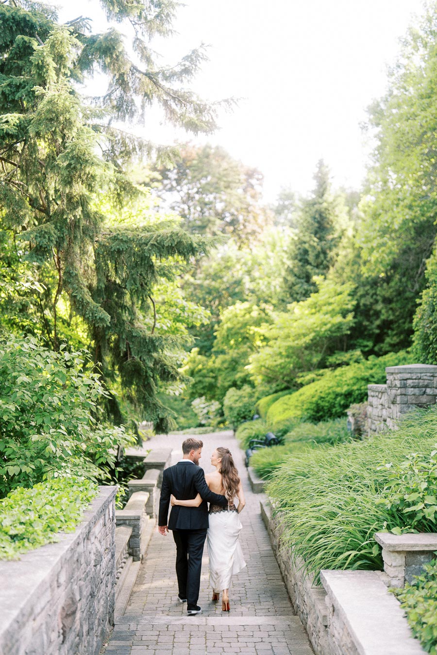 Engaged couple walking arm in arm down a stone path bordered by greenery and stone walls.