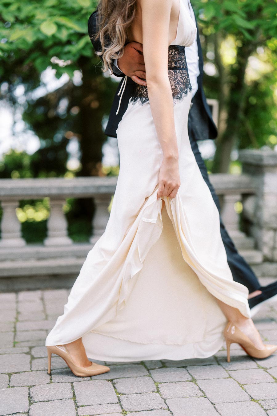 Engaged couple walking on stone path, woman lifts dress, man's arm around her waist.