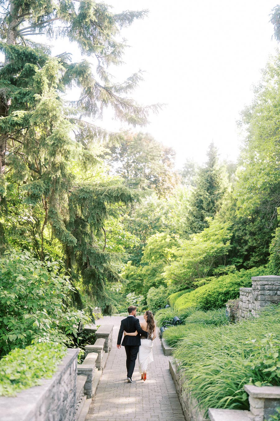 Anastasiia and Petr walking arm-in-arm along a garden stone path bordered by greenery and trees.
