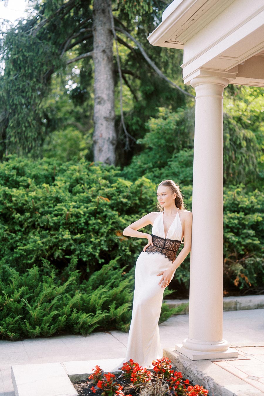 Anastasiia in a long white dress with black lace, leaning on a white column near garden shrubs and red flowers.