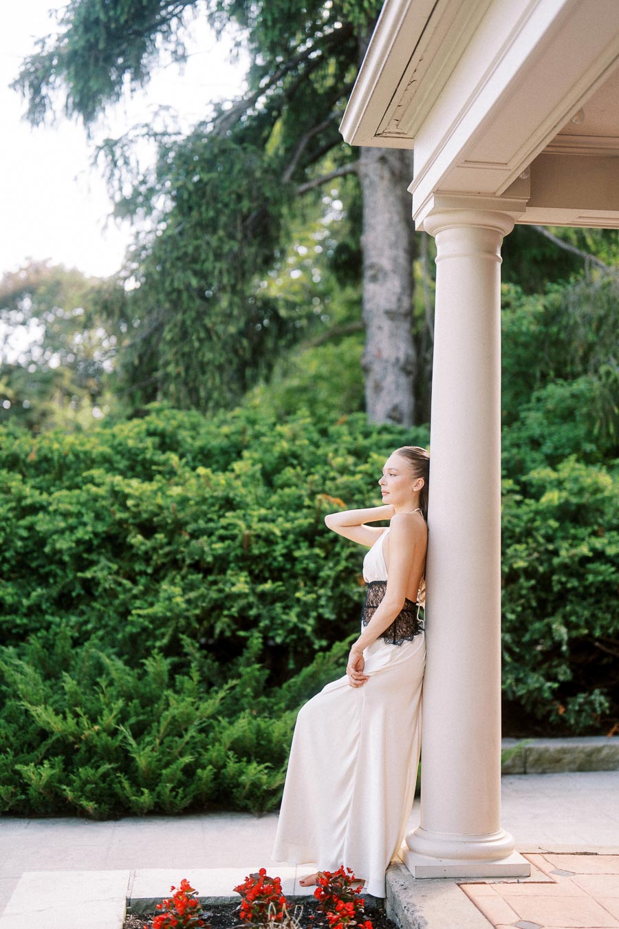 Woman in white dress with black lace leans against white column, surrounded by greenery and red flowers.