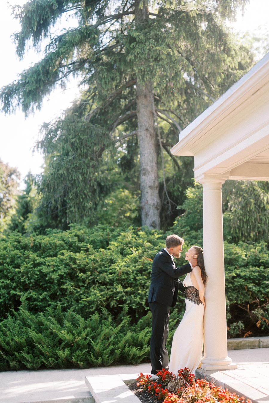 Engaged couple standing by a white column surrounded by greenery and flowers.