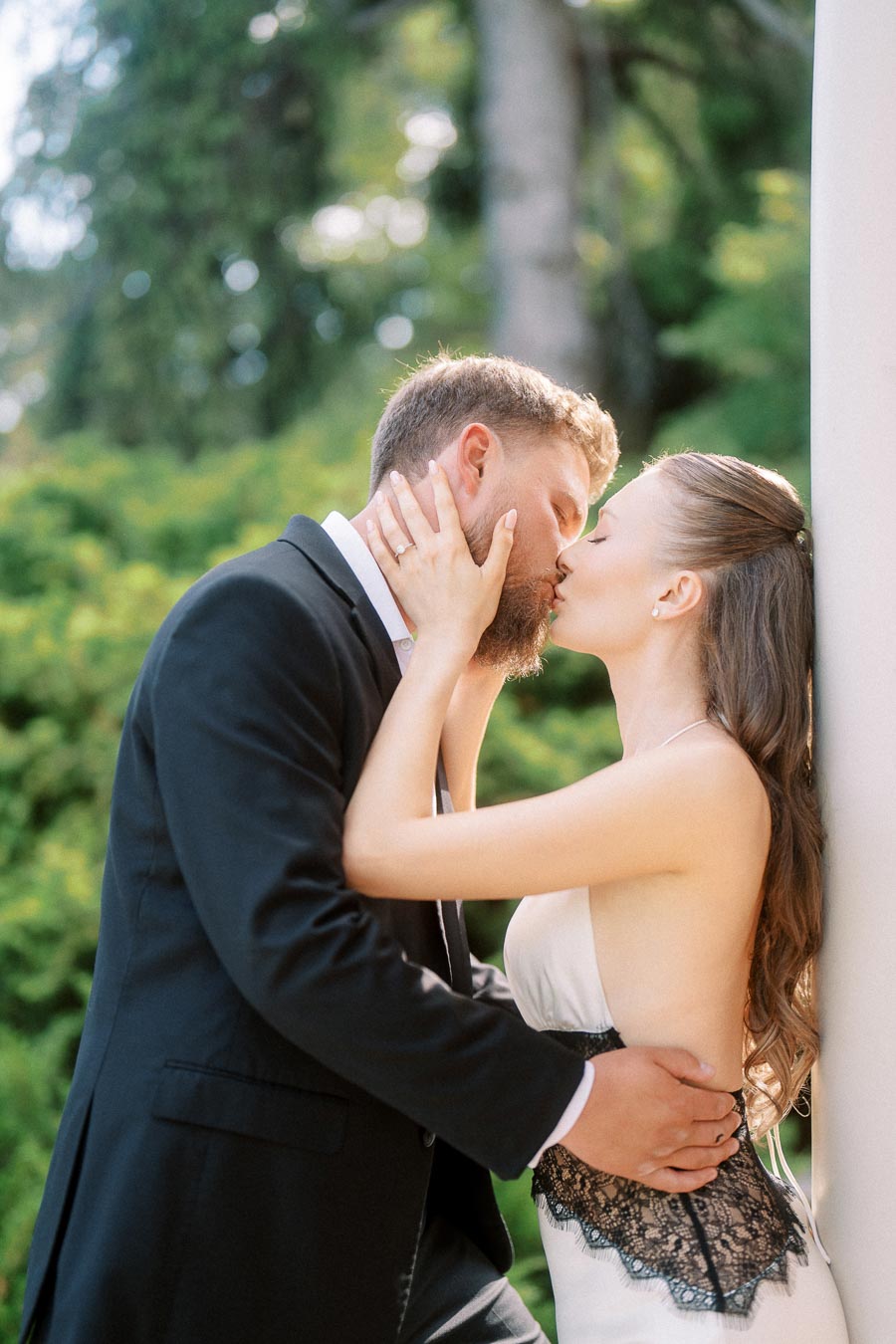 Engaged couple kissing near a white column, greenery in background, engagement ring visible.