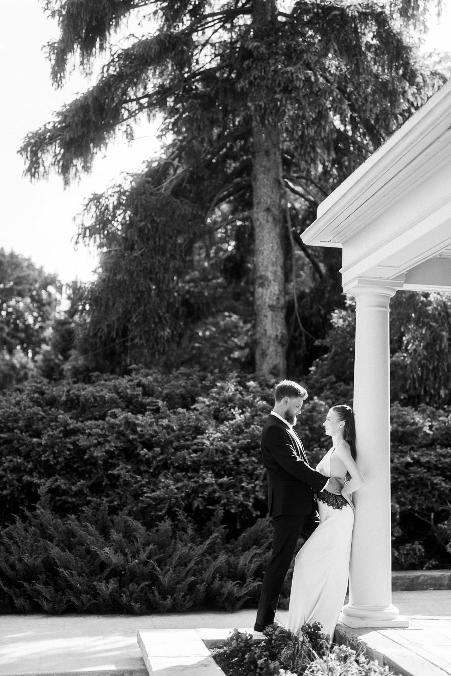 Anastasiia and Petr stand together near a white column, framed by greenery and tall trees in black and white.