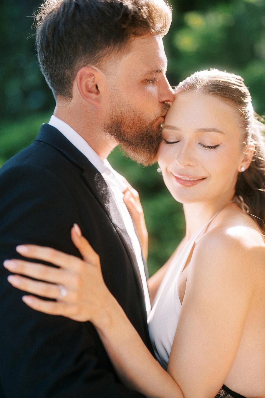 Engaged couple embracing closely, Petr kissing Anastasiia's forehead, sunlight highlighting her engagement ring.