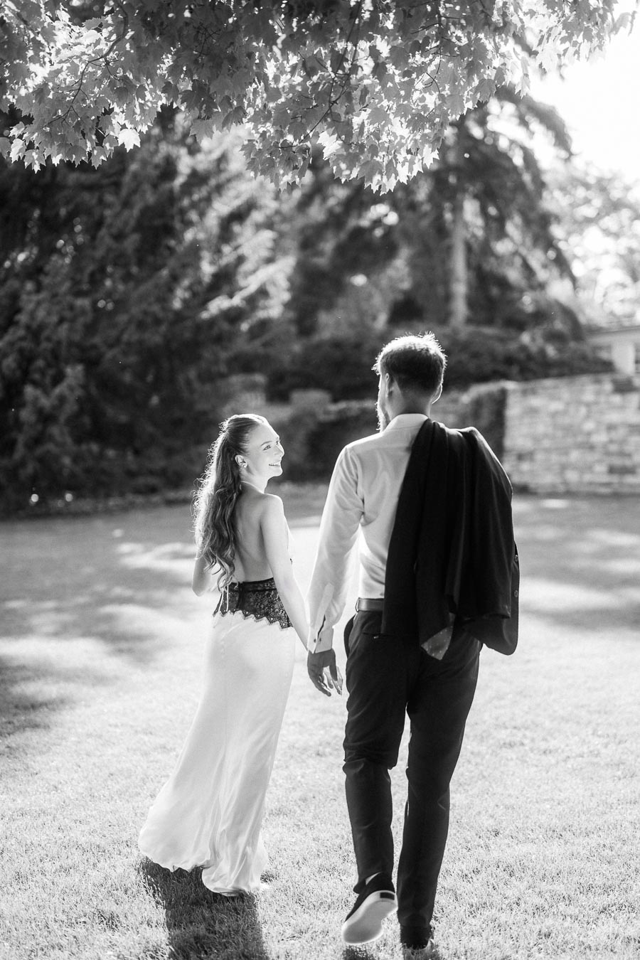 Anastasiia and Petr walking away on grass, holding hands, sunlight streaming through tree branches above.
