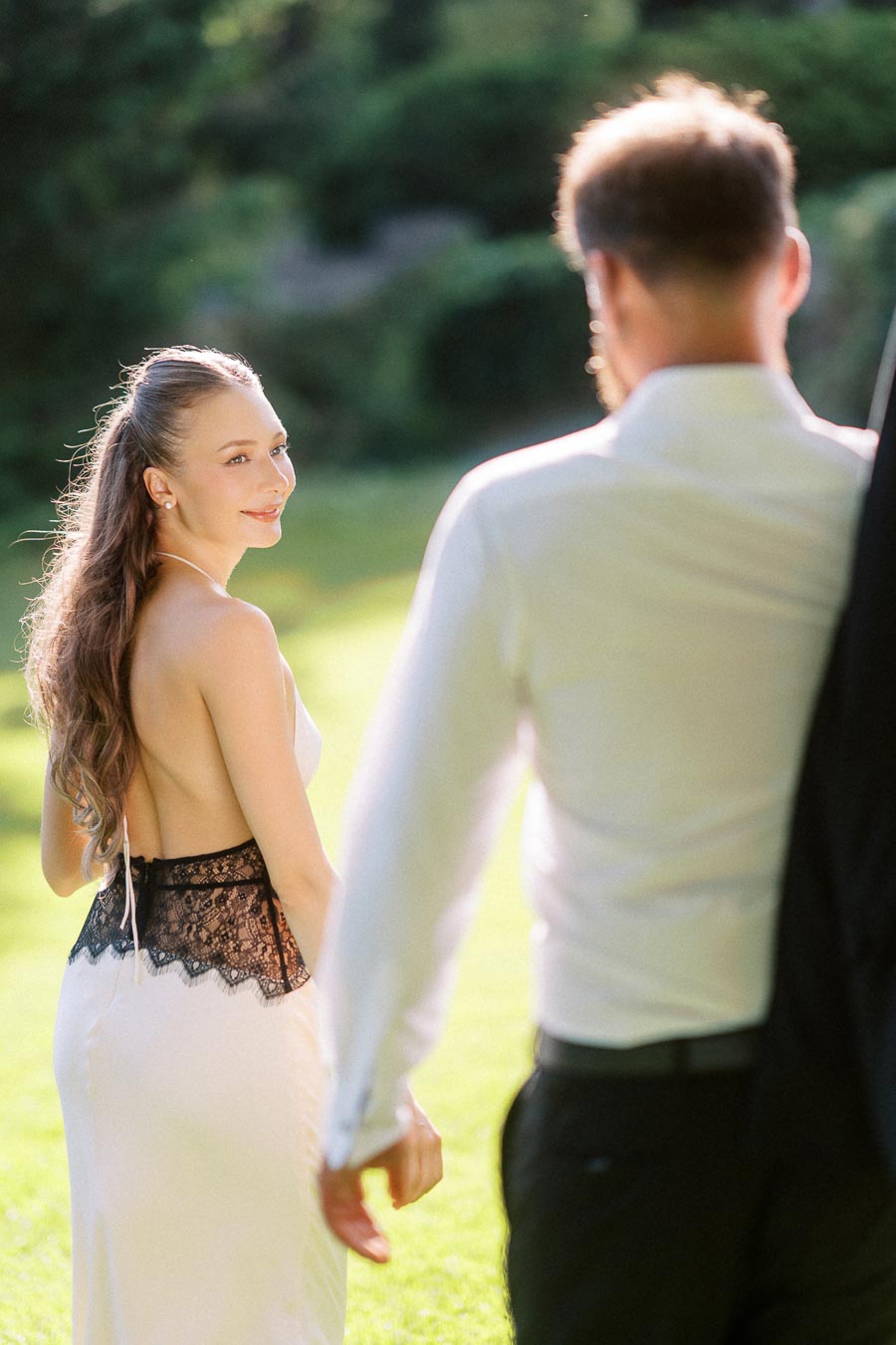 Anastasiia in a white dress with black lace detail turns to look at Petr while walking outdoors on a sunlit lawn.